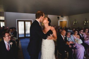 A bride and groom kiss at the altar during a church ceremony, captured by talented documentary wedding photographers. The bride wears a lace gown, the groom a dark suit. Guests in the pews watch the couple in an intimate setting with soft lighting.
