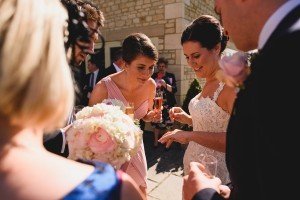 A bride in a white lace dress, surrounded by elegantly dressed people, shows her ring to a woman in a pink dress while Somerset wedding photographers capture the moment. Another woman holds a bouquet of pink and white flowers as they celebrate outdoors in front of a stone building.