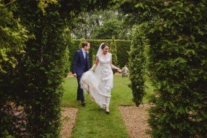 A bride and groom walk through a leafy garden arch in the Cotswolds. The bride wears a lace wedding dress and holds her bouquet, while the groom is in a dark blue suit. They are smiling, surrounded by lush greenery—perfect moments for a wedding photographer to capture.