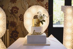 A white three-tiered wedding cake with a gold top tier and floral decoration stands elegantly. The word love is written in gold script on the bottom tier. Large illuminated numbers form a 20 in the background against a floral wallpaper backdrop, perfectly captured by Bristol wedding photographers.