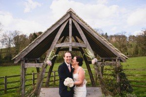 A couple in wedding attire stands under a floral-adorned wooden archway. The bride, holding a bouquet, smiles at the groom in his suit. Captured by talented Bristol wedding photographers, they share this moment in a grassy area with trees and a cloudy sky in the background.