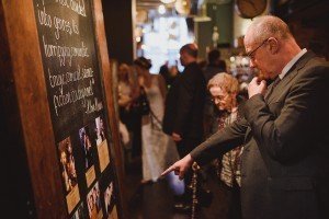 A man in a suit thoughtfully examines a large board displaying photos and text at an elegant event. Nearby, an elderly woman observes the scene. In this dimly lit setting, wedding photographers from Bristol capture the moment as formally dressed guests mingle, including one in a white dress.