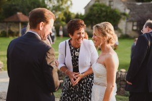 A bride in a white dress smiles as she chats with a man in a suit and a woman in black and white. Set against lush greenery and a stone building, this enchanting garden scene captures the magic moments Somerset wedding photographers dream of preserving.