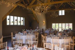 A spacious wedding reception hall in the Cotswolds features exposed wooden beams and chandeliers adorned with string lights. As documented by talented Somerset wedding photographers, round tables with white tablecloths and floral centerpieces sit nearby, with a tiered cake visible in the background.