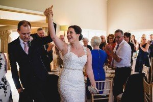 A bride in a lace gown and a groom in a suit hold hands triumphantly as they enter the dining area. Captured by wedding photographers Bristol, guests stand applauding around the decorated tables. The atmosphere is celebratory, with smiling faces and warm lighting.