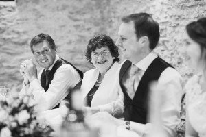 A group of people sitting at a table during a formal event, likely captured by a Bristol wedding photographer. They are wearing formal attire with vests and dresses. The setting appears elegant with a stone wall in the background, and there are flowers on the table. They are smiling and engaged in conversation.