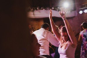 A woman in a pink dress dances with her arms raised in a dimly lit room, surrounded by other people. A bright light shines from above, creating a lively atmosphere captured perfectly by Bristol wedding photographers.