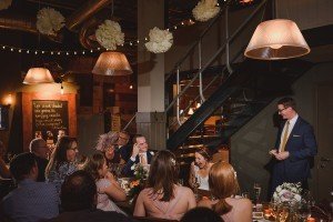 A wedding reception unfolds as a man in a suit stands, speaking warmly to the guests seated at tables. With string lights and paper lanterns casting a golden glow, laughter fills the room. Captured by skilled Bristol wedding photographers, a couple sits together, smiling under the enchanting ambiance.