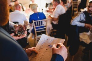 A person is holding a handwritten list while another holds cash and a glass, capturing a candid moment that wedding photographers Bristol would adore. In the background, people are seated at tables, engaged in conversation, creating a lively setting indicative of a social gathering.