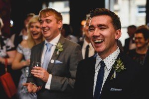A smiling man in a suit with a floral boutonniere and polka dot shirt stands in the foreground, captured by a talented documentary wedding photographer. A man in a gray suit holding a drink and a woman with blonde hair are blurred in the background, adding depth to this joyful moment.