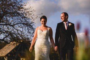 A smiling couple in wedding attire holds hands while walking outdoors, captured beautifully by Somerset wedding photographers. The bride wears a lace gown, and the groom is in a black suit with a pink tie. Behind them, a tree graces the scene under a partly cloudy sky.