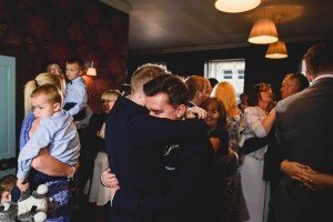 In a warmly-lit room filled with people, two men embrace in a hug. Women holding children and other guests mingle and smile in the background, creating a celebratory atmosphere captured beautifully by documentary wedding photographers.