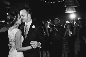 A bride and groom share a dance in a dimly lit venue, surrounded by guests and captured creatively by documentary wedding photographers. The brides lace dress and the groom’s boutonniere shine under string lights, while guests relish capturing the enchanting moment on their phones.