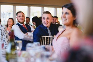 A group of people sitting at a table, dressed in formal attire. A man in a blue shirt and gray tie is looking to the side, smiling. Others around him are also smiling and engaged, captured beautifully by wedding photographers Somerset with a soft focus on the surroundings.