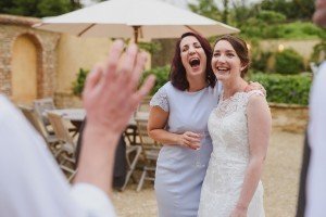 Two women in dresses laugh joyfully outdoors at a wedding captured by talented Bristol wedding photographers. One, in a white lace gown, stands beside the other in a light purple dress holding a glass. They are with others in a garden setting with chairs and a white umbrella.