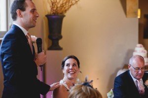 A man in a suit gives a speech with a microphone, standing beside a seated woman in a white dress who smiles up at him, perfectly capturing the candid joy that Bristol wedding photographers are known for. Another suited man looks down at the table as decorative items adorn the backdrop.