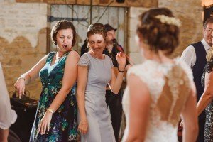 Two women are dancing enthusiastically at an indoor event, with a bride in a lace dress joyfully joining in. Captured by talented Somerset wedding photographers, the scene unfolds against a rustic stone wall as participants revel in the festivities, reflecting the documentary style beautifully.