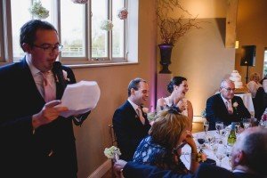 A man in a suit gives a speech at a wedding reception captured by talented Somerset wedding photographers. Seated guests, including the bride and groom, listen and laugh, with floral decorations and a tiered cake adding charm to the scene.