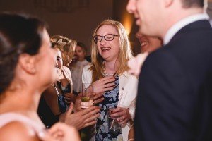 Amidst laughter and lively conversations, a woman in glasses and a floral dress smiles broadly while holding a drink. Shes surrounded by joy, much like the candid moments cherished by documentary wedding photographers capturing authentic emotions at events like this.
