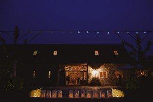 A dimly lit barn at night displays illuminated letters spelling JUST MARRIED at the entrance. Warm light spills from inside, highlighting guests and creating a cozy atmosphere ideal for documentary wedding photographers. String lights decorate above, adding a festive touch.