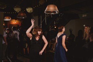 Two women are dancing enthusiastically in a dimly lit room adorned with decorative lights and hanging lamps. In the background, near the bar, a group is either chatting or admiring the dancers—a scene perfect for Somerset wedding photographers to capture such joyful moments.