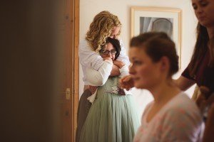 In a warmly lit room, a woman with curly hair lovingly embraces another from behind, exuding happiness and relaxation. In the foreground, slightly out of focus, another woman is seated, enhancing the cozy atmosphere captured perfectly by documentary wedding photographers.