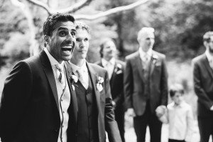 A group of people, including a man with a joyful expression, are dressed in formal suits. They stand outdoors, with trees blurred in the background. A child is also visible in the scene. Captured by wedding photographers from Somerset, this black and white image exudes timeless elegance.