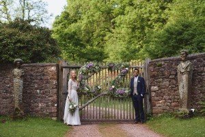 A bride and groom stand beside a rustic gate adorned with a heart-shaped floral arrangement. They are wearing traditional wedding attire. The stone statues flank the gate, and lush greenery surrounds the scene on this bright day, perfectly captured by Somerset wedding photographers.