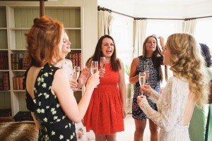 Five women in colorful dresses stand in a living room, smiling and holding champagne glasses. Bookshelves and cream curtains are in the background, capturing a moment even wedding photographers from the Cotswolds would admire. The atmosphere is joyful and celebratory.
