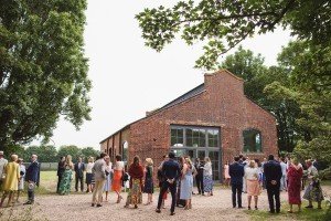 A group of people in formal attire gather outside a brick building with large windows, surrounded by trees and open grass. Capturing this sunny days social gathering, wedding photographers from the Cotswolds blend effortlessly into the scene.