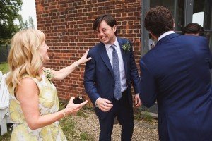 A man in a blue suit and tie laughs while standing outside near a brick wall, perhaps captured by Bristol wedding photographers. A woman in a yellow dress smiles at him, holding a small black box. Two men in suits stand nearby, one facing away from the camera.