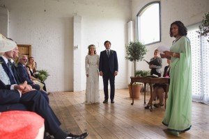 A couple stands holding hands in a well-lit room with a wooden floor and white brick walls, as the officiant in a light green dress reads from a paper. With guests seated to the left and plants decorating the space, its a scene beautifully captured by renowned Bristol wedding photographers.