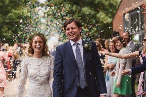 A smiling bride in a white dress and groom in a navy suit walk through a shower of confetti thrown by guests. Captured by Somerset wedding photographers, they are outdoors, surrounded by trees and a brick building, with people celebrating around them.