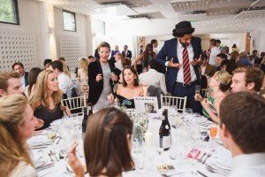 Guests seated around a large round table at an elegant event, where a person wearing a hat stands and gestures expressively, capturing the groups attention. Captured by talented Bristol wedding photographers, glasses and plates adorn the table, as fairy lights twinkle above.