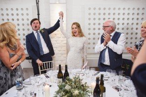 A newlywed couple celebrates at a wedding reception, with wedding photographers from Bristol capturing each triumphant moment. The bride and groom hold hands, surrounded by smiling guests clapping. The table is set with wine bottles, glasses, and floral decorations. The mood is joyful and festive.
