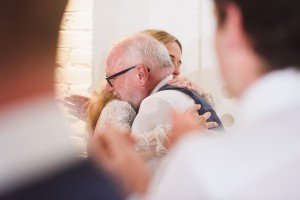 An elderly man with glasses and a white beard warmly hugs a woman, possibly at a celebration. Both are in formal attire, their embrace captured with the gentle focus of wedding photographers from Bristol. In the foreground, two blurred figures suggest a larger gathering or event.