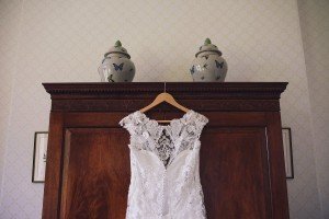 A white lace wedding dress hangs on a wooden wardrobe, captured beautifully by Somerset wedding photographers. Two decorative vases with butterfly designs sit gracefully on top, against a backdrop of patterned wallpaper adorned with framed pictures.