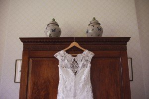 A white lace wedding dress hangs on a wooden wardrobe, its intricate bodice detailing catching the light. Two decorative ceramic jars with butterfly designs are displayed on top, available for wedding photographers Bristol to capture. The wallpaper features a subtle diamond pattern.