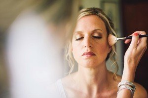 A woman with closed eyes is having makeup applied with a brush, her light blonde hair elegantly styled in an updo. Shes wearing a white top against a softly blurred background, perfect for capturing by Somerset wedding photographers.
