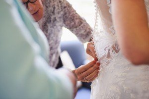 Close-up of a brides intricate lace wedding dress being buttoned by two people. One person’s hands focus on fastening the delicate buttons along the back, capturing a candid moment that documentary wedding photographers in Somerset would treasure.
