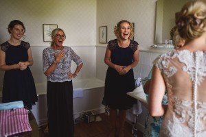 A bride in a lace gown stands with her back to the camera, showing her dress to three women in a bathroom. Captured by a Bristol wedding photographer, the women react with joy and excitement, wearing navy dresses and a lace top with a black skirt. The room features light walls and framed art.