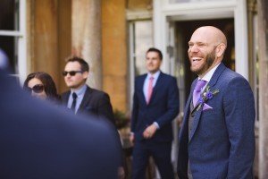 At a lively social gathering, a bald man in a blue suit and purple tie laughs amidst three others in suits, one wearing sunglasses. They stand before a rustic building with tall columns. This moment captures the candid charm that Somerset wedding photographers are renowned for.