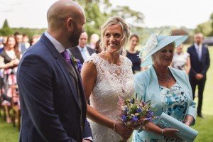 A bride in a lace gown holds a bouquet and smiles at a bald man in a suit during an outdoor ceremony, perfectly captured by Somerset wedding photographers. An older woman in a floral dress and blue hat stands beside them, while guests gather on the grassy field in the background.