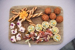 A wooden platter showcases a delightful array: breadsticks, falafel balls, petite pastry cups with blue cheese and herbs, oat crackers topped with cream cheese and flowers, and skewers of cured meat and olives, all garnished with an orange flower—perfect for a charming Cotswolds wedding feast.
