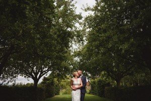 A couple embracing in a lush garden, perfectly captured by skilled wedding photographers from Somerset. They stand on a grassy path framed by tall trees with dense, leafy branches. The woman in her white lace gown and the man in his dark suit are bathed in dappled sunlight filtering through the leaves.