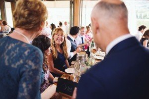 Amidst the lively celebration, captured as if by documentary wedding photographers, a woman in a blue dress smiles brightly across the table. Plates, glasses, and bottles adorn the table, enhancing the festive atmosphere shared by all around her.