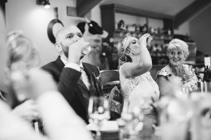 A bride and groom are seated at a table, raising glasses in a celebratory toast. The bride sips from a glass, while the groom drinks from a bottle. An older woman smiles nearby. Captured by talented Somerset wedding photographers, the scene is warmly lit and cheerful, with bottles and glasses on the table.
