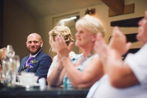 A wedding scene with a man in a suit and purple tie smiling, seated at a table. People around him are clapping. The focus is on the man and a woman applauding in front. Captured by skilled documentary wedding photographers, the background reveals a tastefully blurred interior.