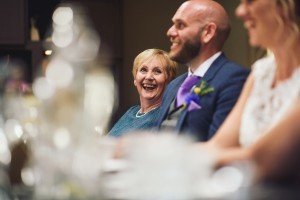 Three people sit at a table, smiling and laughing like subjects in a lively documentary wedding. The focus is on an older woman with short blonde hair and a blue top. Beside her, a bald man in a suit with a purple tie and a woman in white share the moment—a delightful scene captured by Bristol wedding photographers.