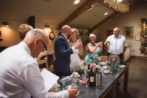 A group of people gathered around a table, dressed in formal attire, raising their glasses in a toast. Captured by documentary wedding photographers, the indoor celebration is adorned with drinks and decorations, preserving cherished memories in each candid frame.